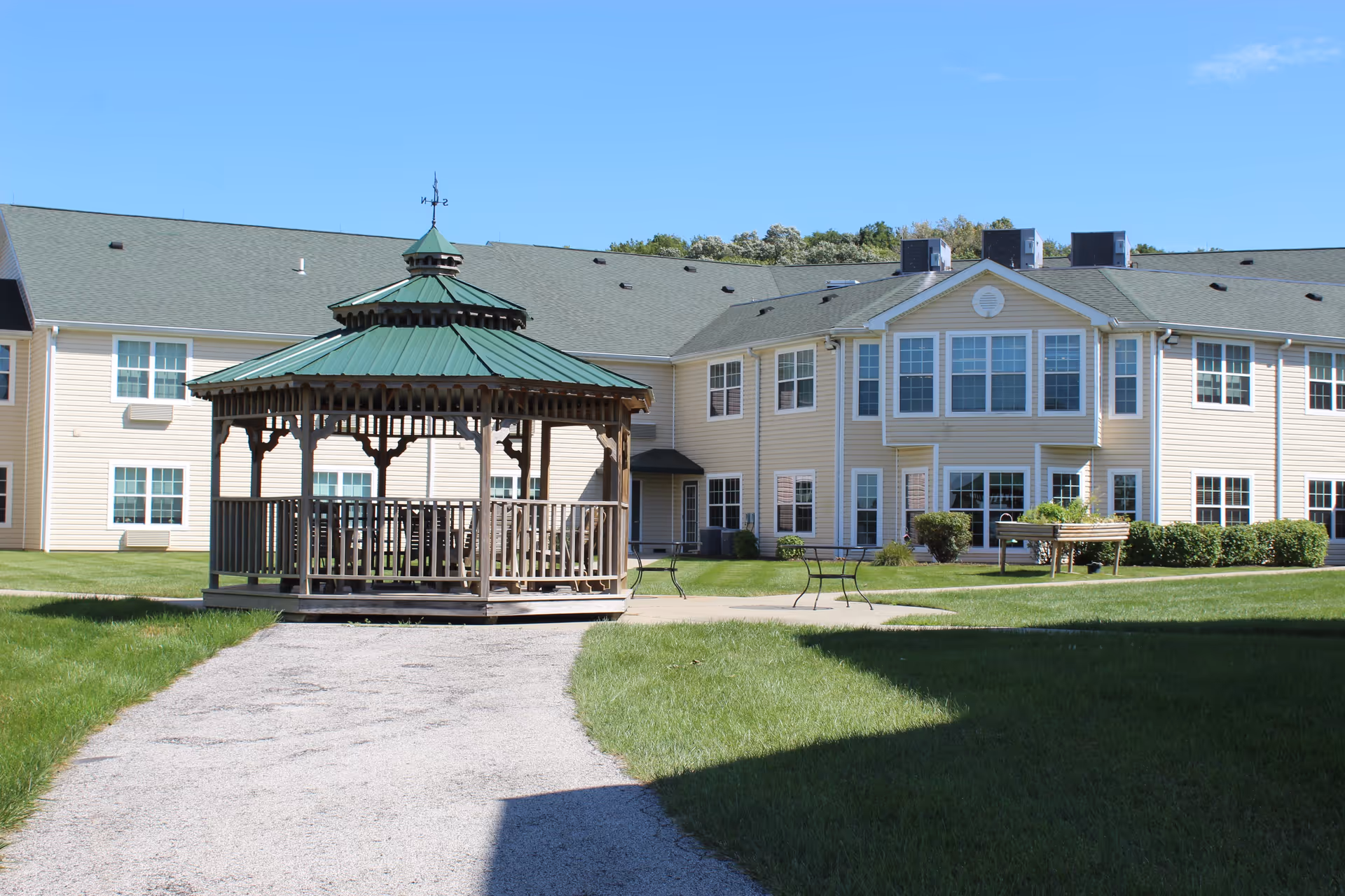 Outdoor view of Knollwood Retirement Community featuring a wooden gazebo with a green roof on a gravel path, surrounded by green grass and a beige two-story building with many windows under a clear blue sky.