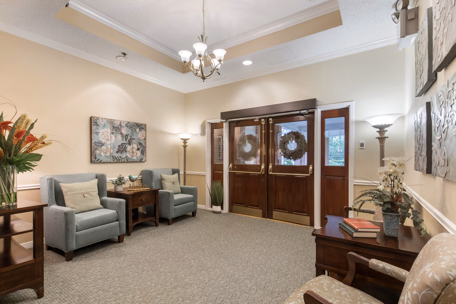 A cozy and well-lit sitting area near a set of double wooden doors with glass panels decorated with wreaths. The room features two light blue armchairs with cushions, a wooden side table with a small plant and decorative bird, a floor lamp, and a wooden table with books and a potted plant. The walls are painted beige and adorned with floral artwork and decorative panels.