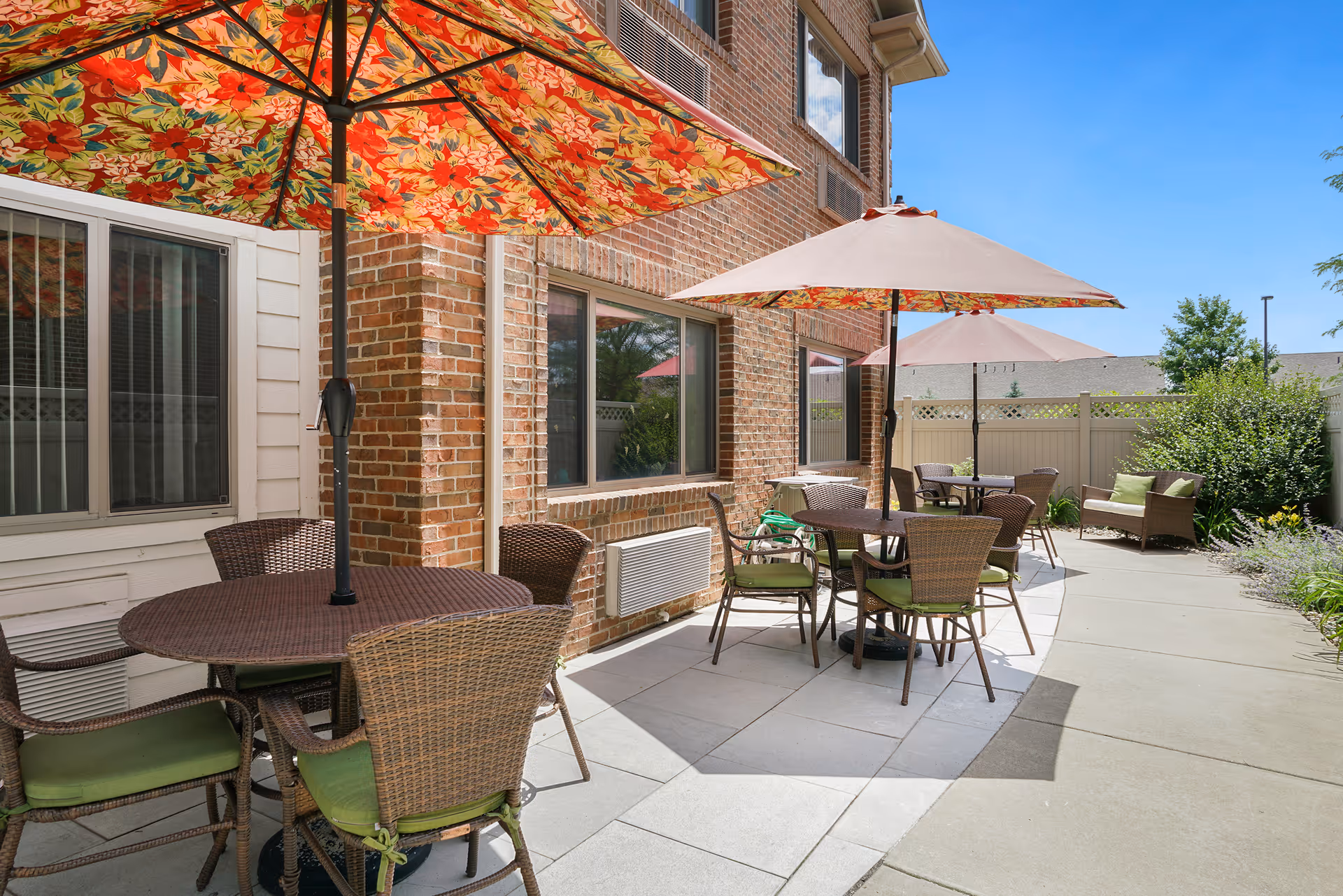Outdoor patio area with several round tables and wicker chairs with green cushions. Each table has a large umbrella, some with floral patterns and others plain. The patio is adjacent to a brick building with windows, and there is a white privacy fence and greenery in the background under a clear blue sky.