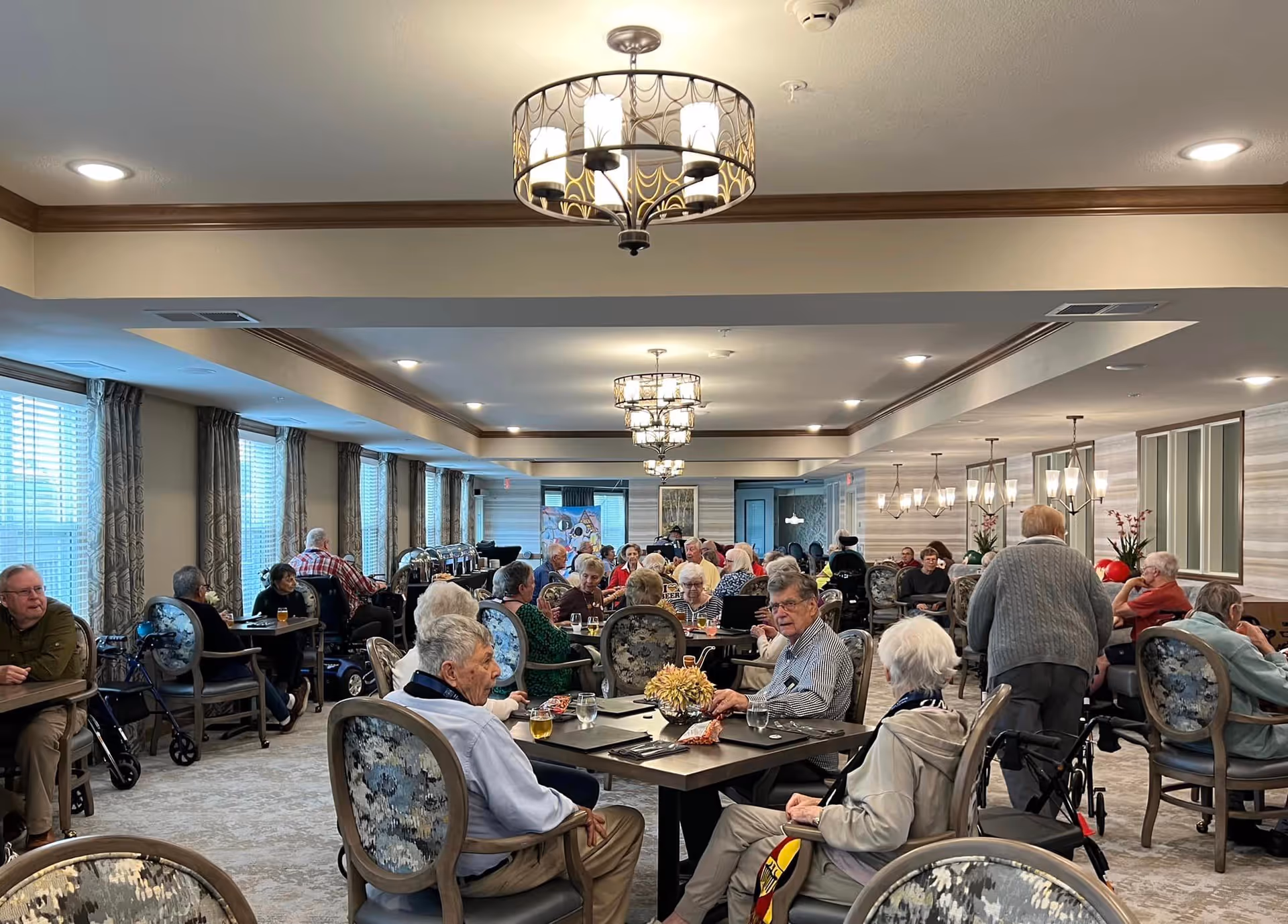 A large dining room filled with elderly residents sitting at tables, eating and socializing. The room has multiple chandeliers, large windows with curtains, and a warm, inviting atmosphere.
