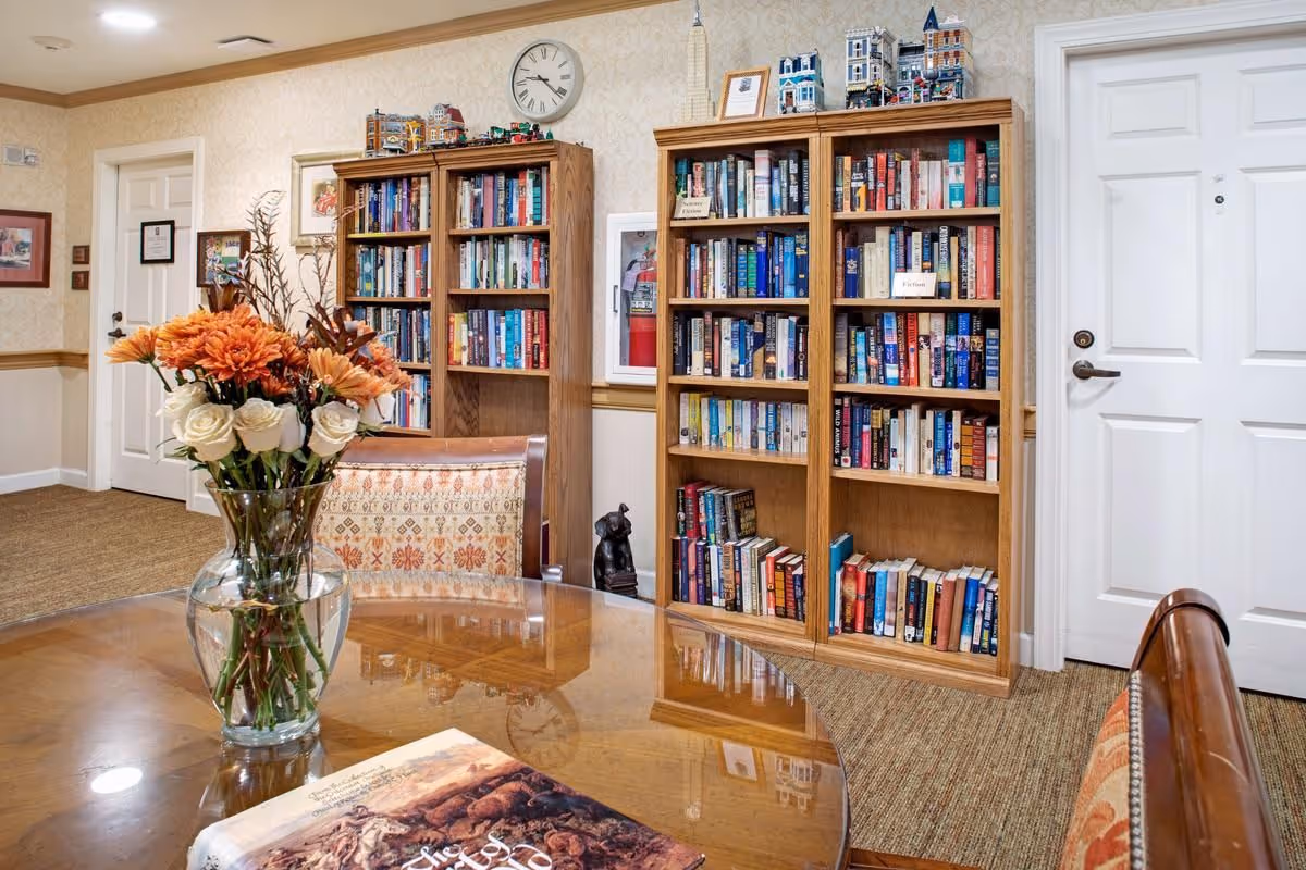 A cozy common room with wooden bookshelves, a round table topped by a vase of flowers, and upholstered chairs.