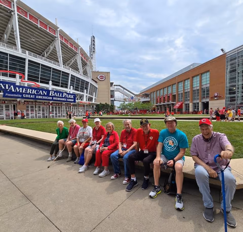 A group of elderly people sitting on a low concrete wall outside Great American Ball Park. They are dressed casually, some wearing red clothing and baseball caps, enjoying a day out near the stadium with a grassy area behind them and the ballpark structure visible in the background.