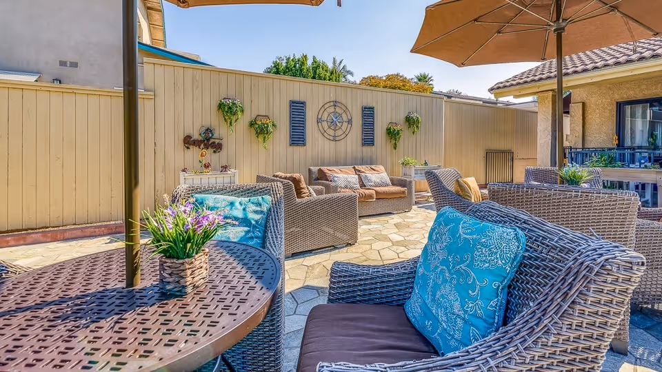 Sunny outdoor patio with wicker seating, blue patterned cushions, umbrellas, potted plants, and a decorative wooden fence.