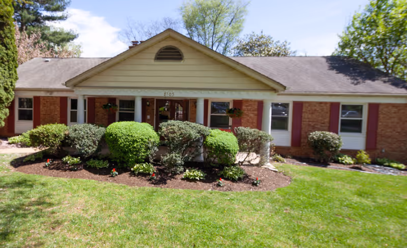 Single-story brick building with a gabled roof and white columns at the entrance, surrounded by neatly trimmed bushes and a green lawn under a clear blue sky.