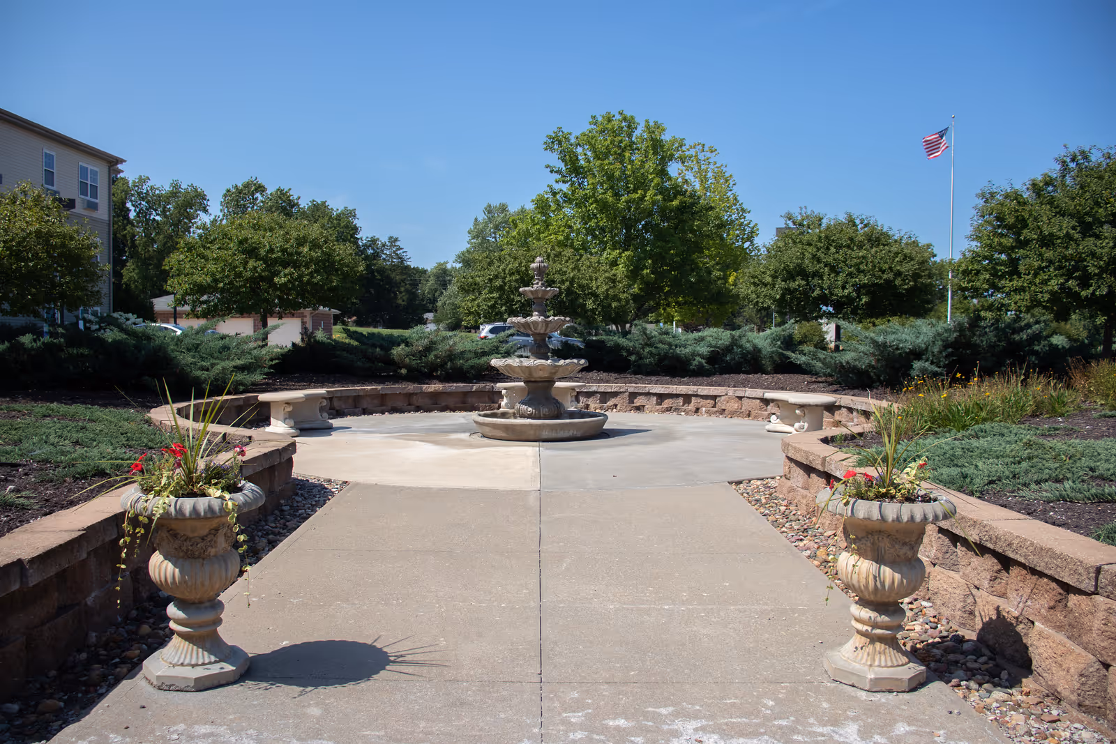 Outdoor garden area with a three-tiered stone fountain in the center, surrounded by a circular stone bench. Two decorative stone planters with flowers flank the concrete pathway leading to the fountain. Trees and shrubs are visible in the background under a clear blue sky, with an American flag on a flagpole to the right.