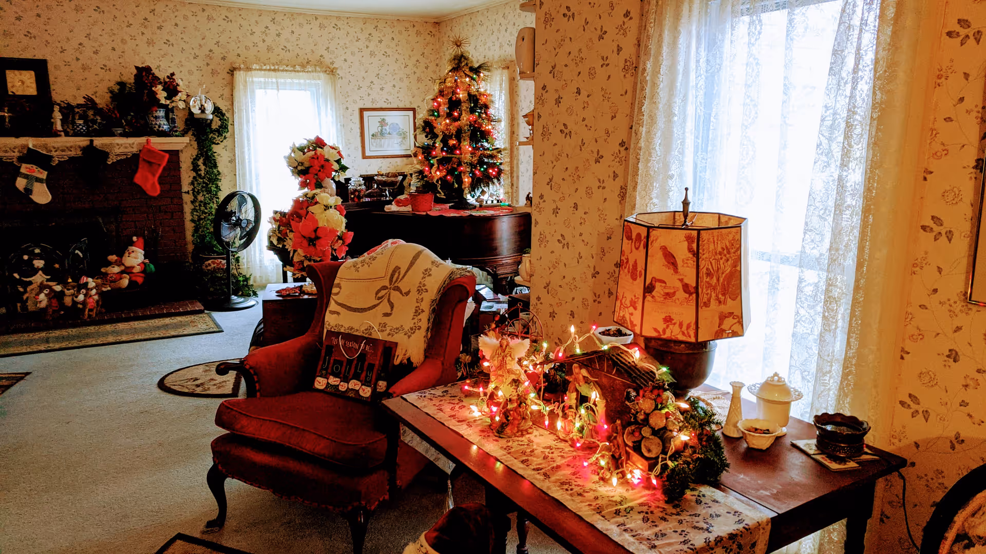 A cozy living room decorated for Christmas with a red armchair draped with a white throw, a wooden table adorned with Christmas lights and decorations, a lamp with a patterned shade, and a window with lace curtains. In the background, there is a decorated Christmas tree, a fireplace with stockings and holiday figurines, and floral wallpaper covering the walls.