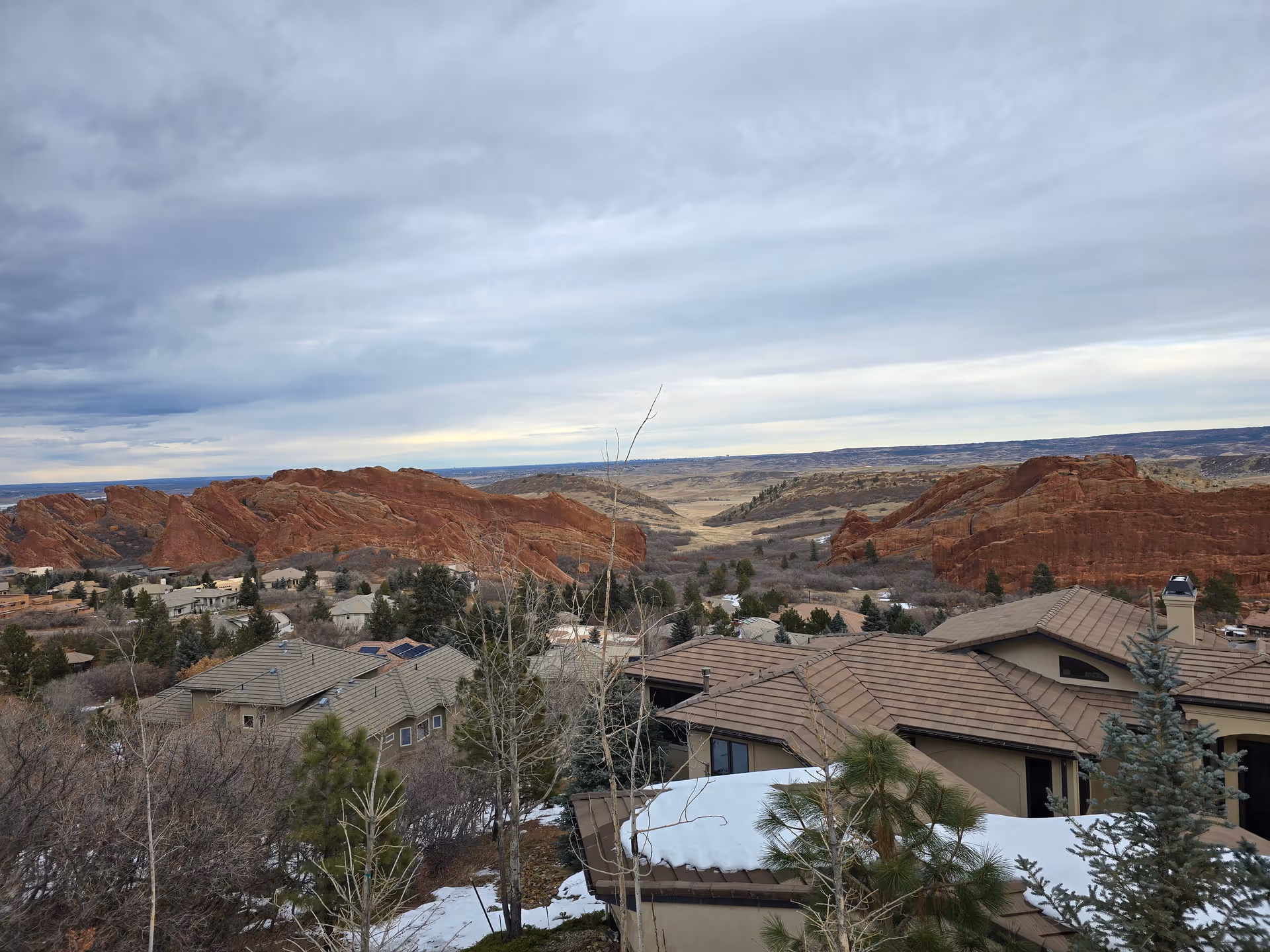 View of a residential area with houses featuring brown tiled roofs surrounded by trees, with large red rock formations and a vast landscape under a cloudy sky in the background.