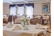 Bright dining room with tables set with folded napkins and floral centerpieces, upholstered chairs, and windows with valances.