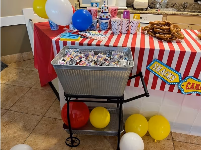 A festive snack table with a red and white striped tablecloth, decorated with colorful balloons in red, yellow, white, and blue. The table holds pretzels, popcorn containers, cups, napkins, and a metal tub filled with juice pouches. The setting appears to be indoors with tiled flooring and a kitchen counter in the background.