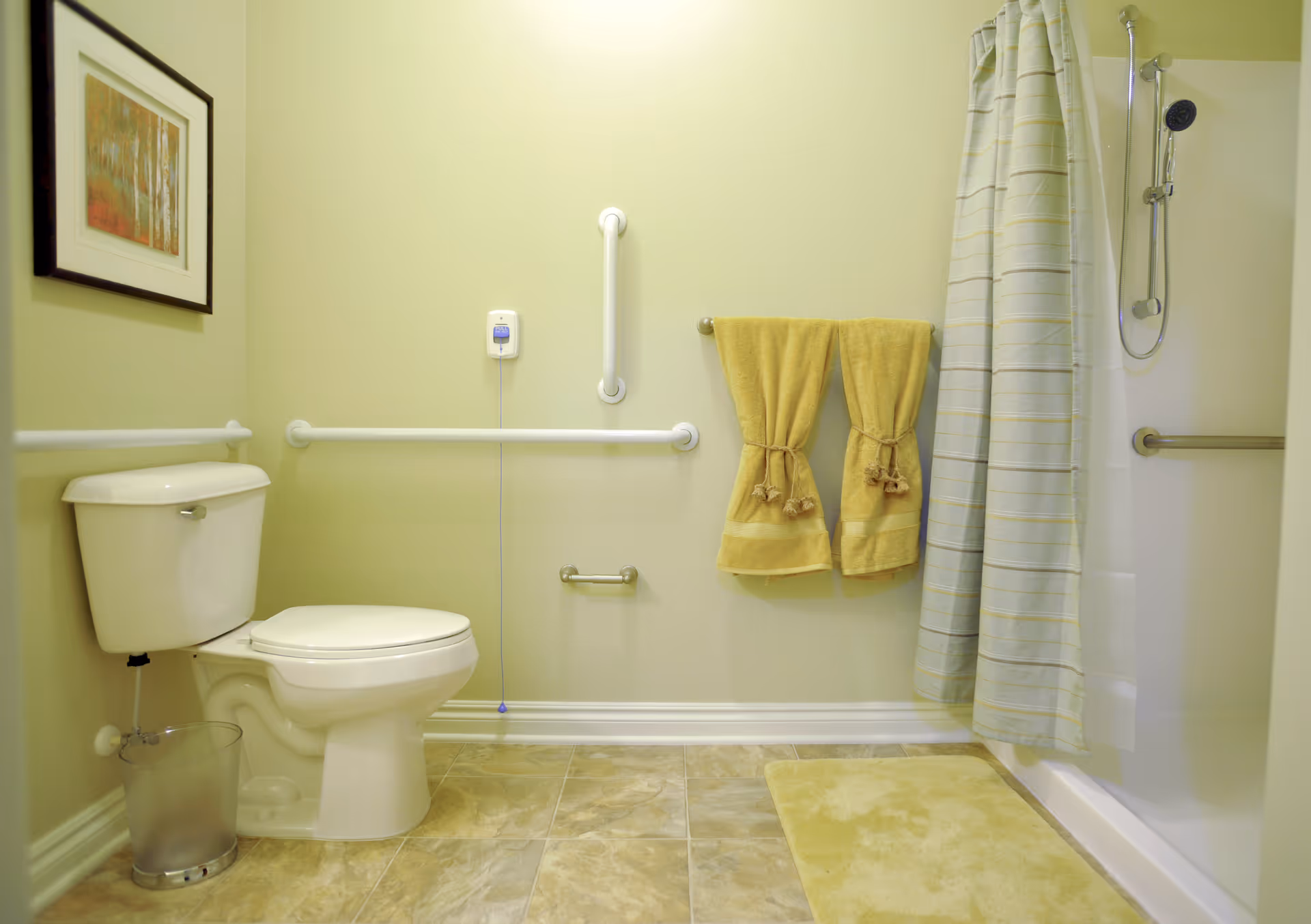 A clean and accessible bathroom with a white toilet, grab bars on the walls, a shower with a striped curtain, two yellow towels hanging on a towel rack, a yellow bath mat, a small trash can, and a framed picture on the wall.