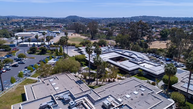 Aerial view of a senior living facility with multiple flat-roofed buildings surrounded by trees and parking lots, set against a backdrop of a suburban area with hills in the distance under a clear blue sky.