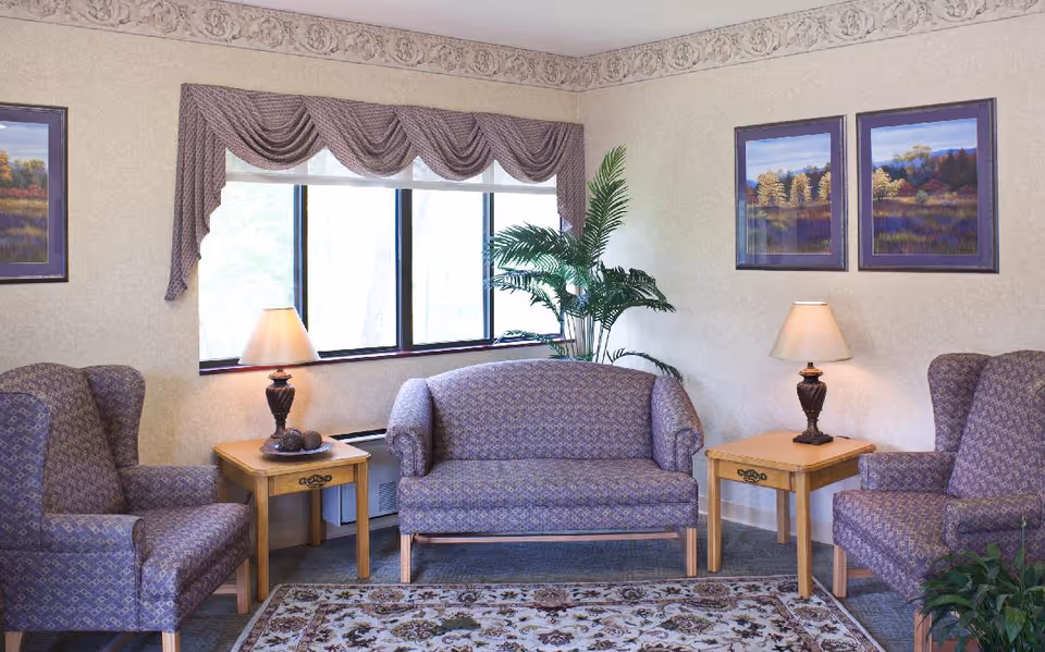 A cozy living room area with two patterned armchairs and a matching loveseat arranged around two wooden side tables with lamps. The room has a large window with decorative valances, two framed landscape paintings on the wall, a patterned area rug on the floor, and a tall green plant in the corner.
