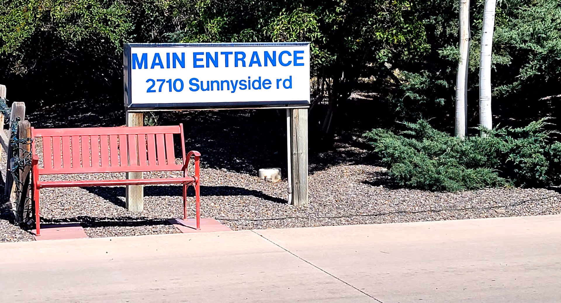 Outdoor main entrance sign reading 'MAIN ENTRANCE 2710 Sunnyside rd' beside a red bench and landscaping.