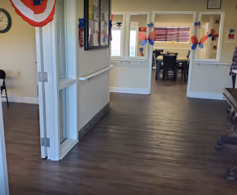 Interior hallway with wood flooring leading to a room with a dining table and chairs. The walls are decorated with patriotic red, white, and blue decorations including butterflies and a bunting. There is a clock on the wall and a bulletin board with papers.