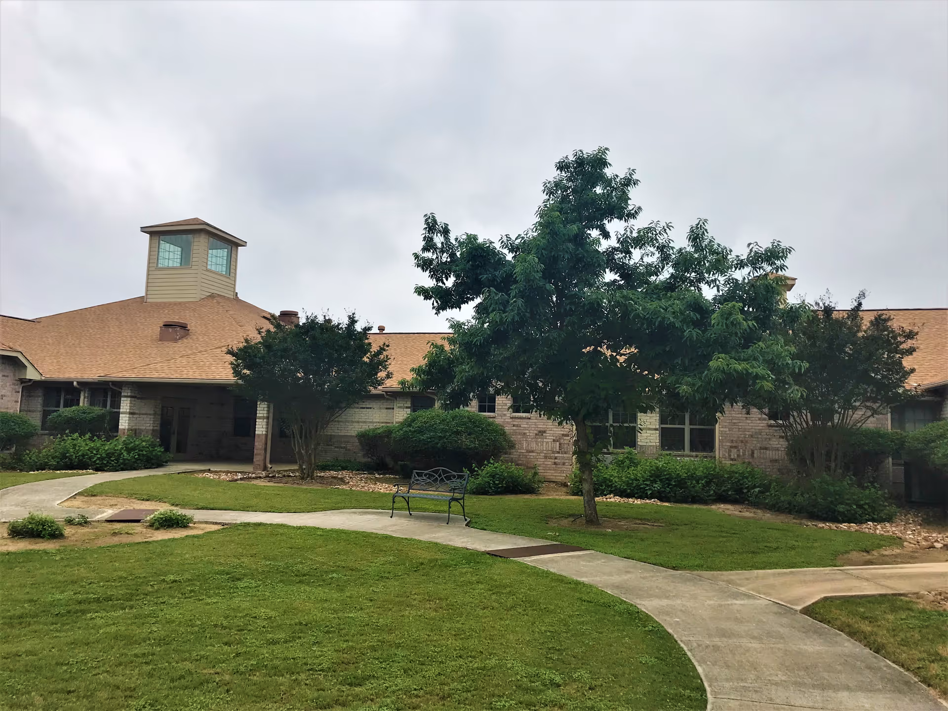 Exterior view of a single-story brick building with a tan roof and a small tower-like structure on top. The building is surrounded by green grass, trees, and shrubs, with a curved concrete walkway leading to the entrance. There is a metal bench near the walkway.