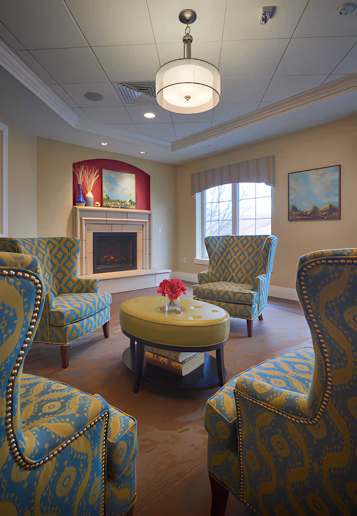 A cozy living room area with four patterned armchairs arranged around a round ottoman with a small vase of red flowers on top. There is a fireplace with decorative vases and a painting above it, a window with a valance, and another painting on the wall. The ceiling has recessed lighting and a hanging light fixture.