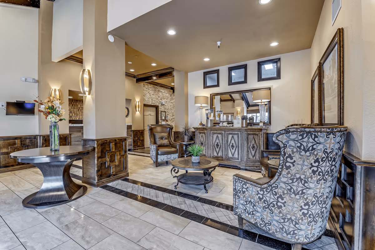 A well-lit interior lounge area with patterned armchairs, a round coffee table with a small plant, a wooden sideboard with lamps and decorative items, and a round table with a vase of flowers. The space features tiled floors, beige walls, and recessed lighting.