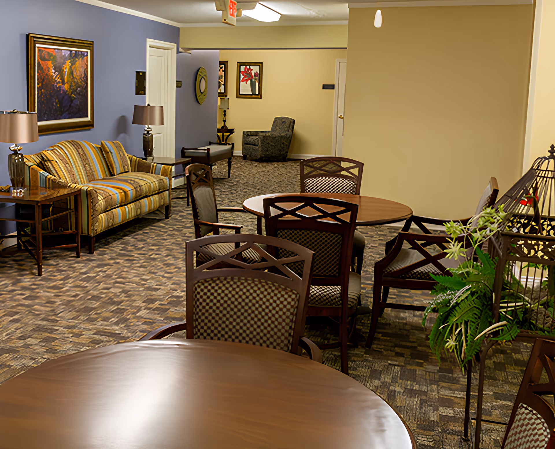 Interior common area of a senior living facility featuring round wooden tables with patterned chairs, a striped sofa with side tables and lamps, framed artwork on the walls, and a carpeted floor. There is also a decorative plant stand with greenery on the right side.