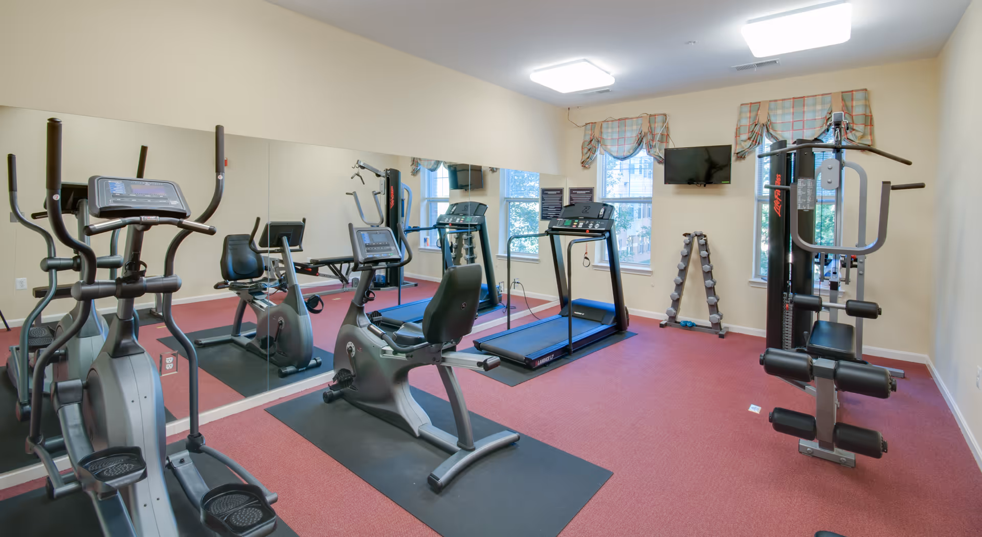 A senior living facility fitness room with exercise equipment including an elliptical machine, a recumbent bike, a treadmill, a multi-function weight machine, and a rack of dumbbells. The room has red carpet flooring, beige walls, two windows with plaid valances, a wall-mounted TV, and a large mirror covering one wall.