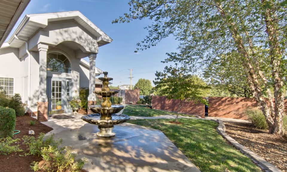 Outdoor view of a senior living facility entrance with a multi-tiered water fountain in the foreground, surrounded by greenery, trees, and a brick wall under a clear blue sky.