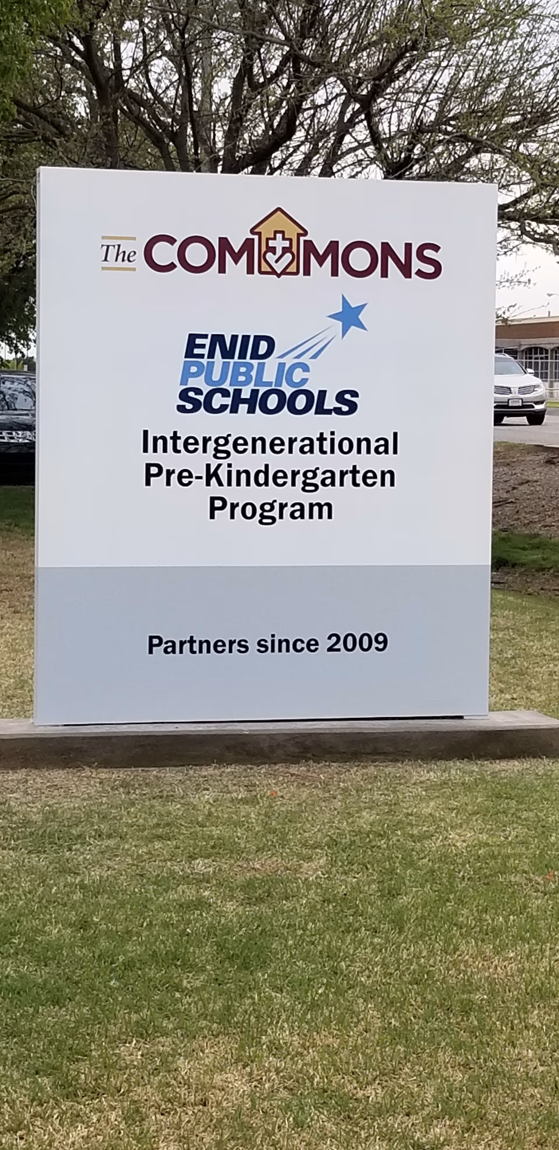 Outdoor sign for The Commons and Enid Public Schools Intergenerational Pre-Kindergarten Program, stating they have been partners since 2009, with grass and trees in the background.