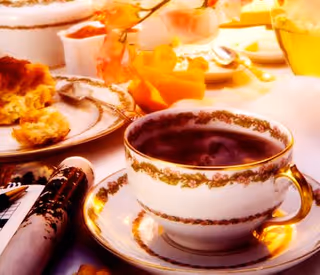 A close-up view of a tea cup filled with tea on a matching saucer, surrounded by plates with pastries and a flower decoration on a table set for tea.