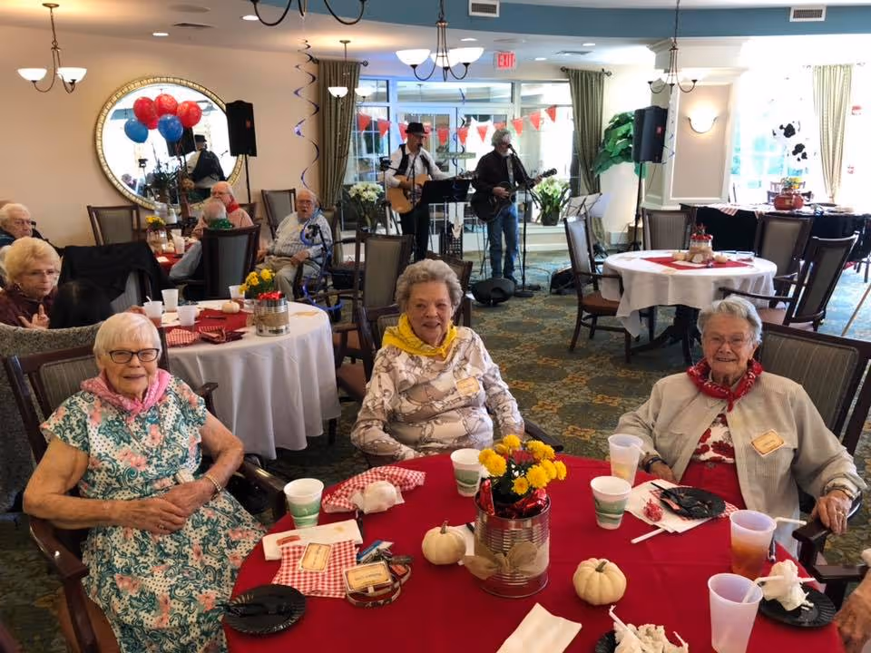 Three elderly women sitting around a table with a red tablecloth, decorated with small white pumpkins and a flower centerpiece. They are in a communal dining area with other seniors seated at tables in the background. Two musicians are performing with guitars near the entrance, and festive balloons and banners decorate the room.