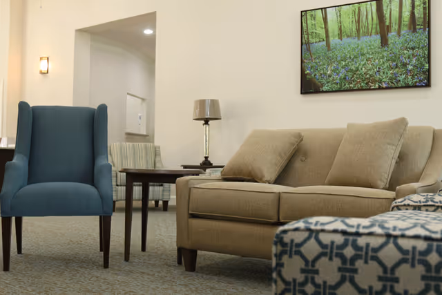 A cozy living room area with a beige sofa adorned with two matching cushions, a blue armchair, a patterned ottoman, a wooden side table with a lamp, and a framed picture of a forest with blue flowers on the wall.