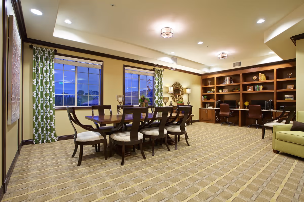 A spacious, well-lit room with a large wooden dining table surrounded by eight chairs with cushioned seats. The room features two large windows with green and white patterned curtains, a built-in wooden bookshelf and desk area with three office chairs, decorative lamps, and a green armchair in the corner. The carpet has a beige and light brown striped pattern, and the walls are painted a soft yellow with dark wood trim.