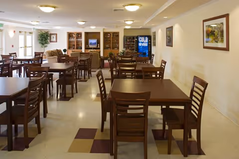 A spacious dining area with multiple wooden tables and chairs arranged neatly. The room is well-lit with ceiling lights and decorated with framed artwork on the walls. In the background, there is a vending machine and a seating area with a couch and a television.