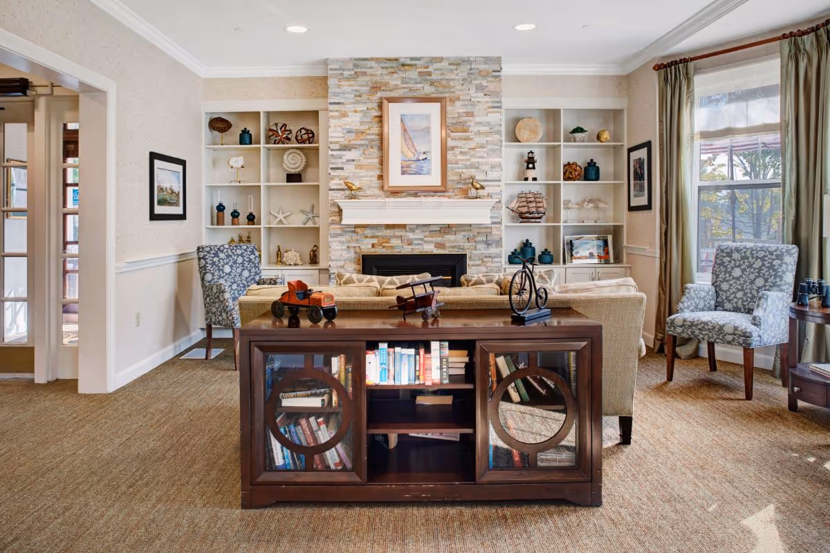 A cozy living room with a stone fireplace centered on the back wall, flanked by built-in white shelves decorated with various ornaments and books. In front of the fireplace is a beige sofa with a dark wooden cabinet behind it, displaying books and decorative items on top. Two patterned armchairs are positioned on either side near windows with green curtains, allowing natural light to fill the room.