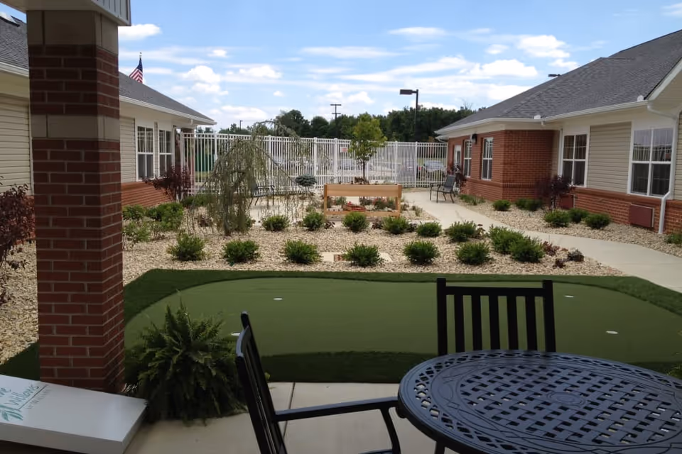 Outdoor courtyard area at The Willows at Willard featuring a small putting green, patio table and chairs, landscaped garden beds with shrubs and small trees, and surrounding single-story buildings with brick and siding exterior under a partly cloudy sky.