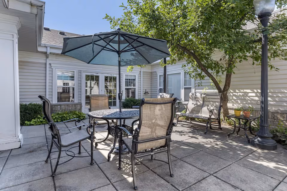 Outdoor patio area with a round metal table and four chairs, shaded by a large blue umbrella. There is a tree providing additional shade, a bench with cushions, a small side table with potted plants, and a tall streetlamp-style light fixture. The patio is surrounded by light-colored siding walls with multiple windows and a glass door.