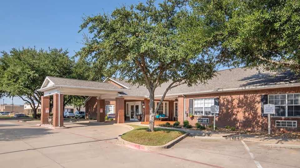 Exterior view of a single-story brick building with a covered entrance supported by brick columns. There are trees and a small landscaped area with grass and shrubs near the entrance. The sky is clear and blue.