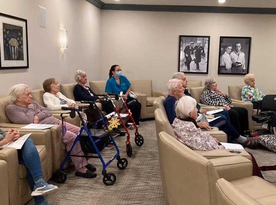 A group of elderly individuals seated in a common room with beige armchairs and sofas, attentively facing forward. One caregiver wearing a blue uniform and a face mask is seated among them. The room has neutral-colored walls adorned with black and white framed photos, and the floor is carpeted.