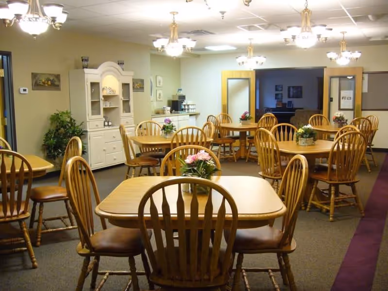 A dining room with multiple wooden tables and chairs arranged neatly. Each table has a small floral centerpiece. The room has beige walls, carpeted floors, and ceiling lights with chandeliers. There is a white cabinet and a counter with a coffee machine along one wall.