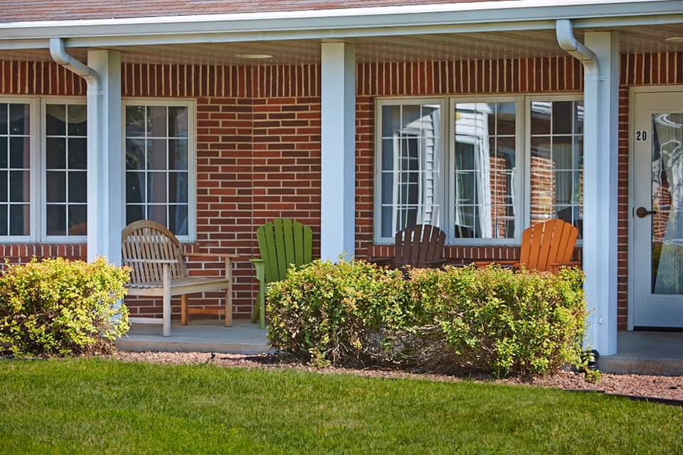 Brick-front porch with several wooden chairs, windows, a door, and trimmed shrubs.