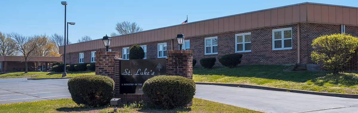 Exterior view of Saint Luke's Nursing Center, a single-story brick building with multiple windows, surrounded by trimmed bushes and a parking lot under a clear blue sky.