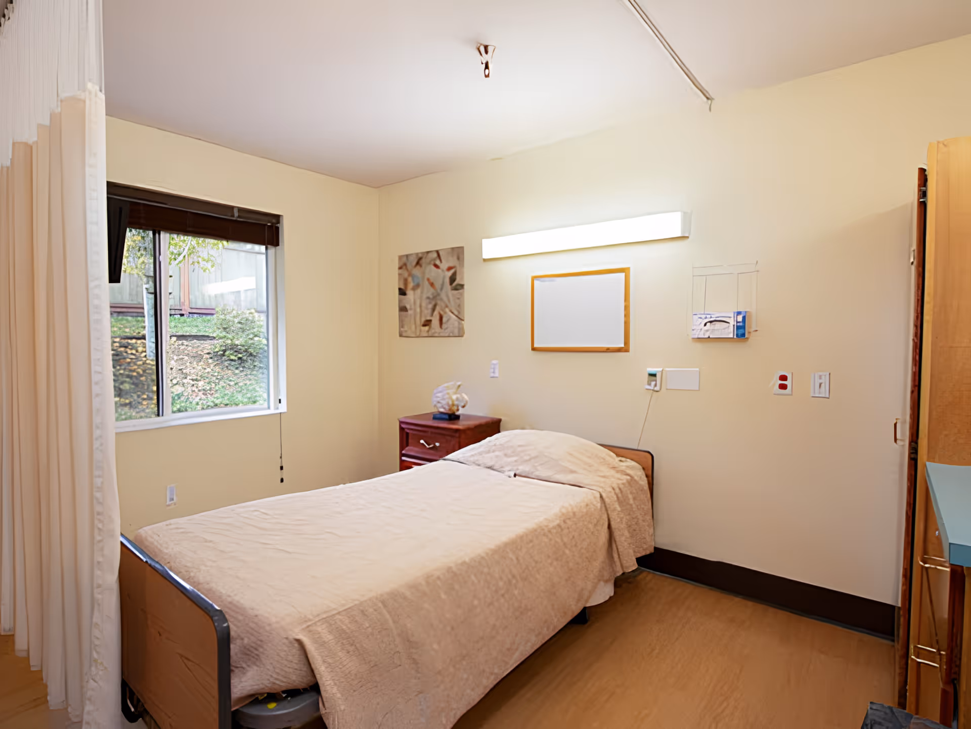 A simple bedroom in a senior living facility with a single bed covered in a beige quilt, a wooden nightstand with a decorative item, a window with a view of greenery outside, a wall-mounted light fixture above the bed, and a curtain partially visible on the left side.