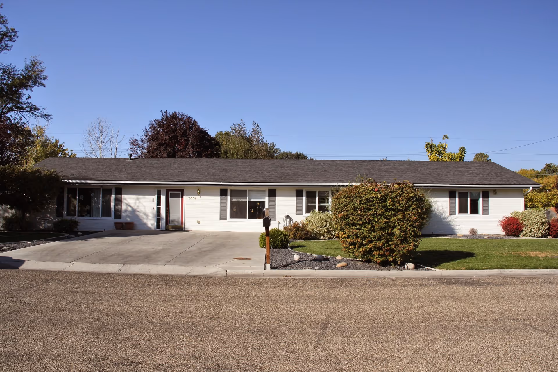 Single-story white building with a dark roof, surrounded by a well-maintained lawn and bushes, under a clear blue sky.