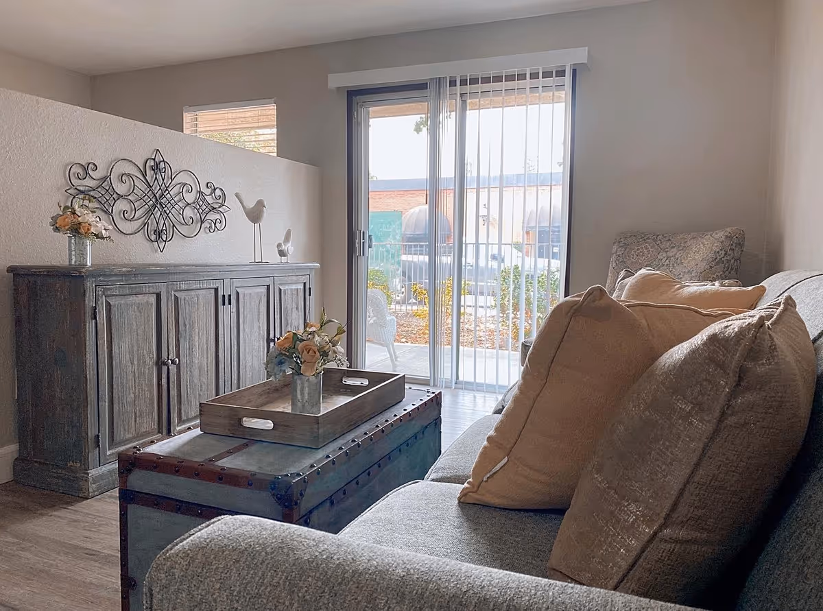 A cozy living room with a gray couch adorned with beige and brown pillows, a wooden chest used as a coffee table with a tray and flower arrangement on top, a rustic wooden cabinet with decorative items including flowers and bird figurines, and a sliding glass door with vertical blinds leading to an outdoor patio area.
