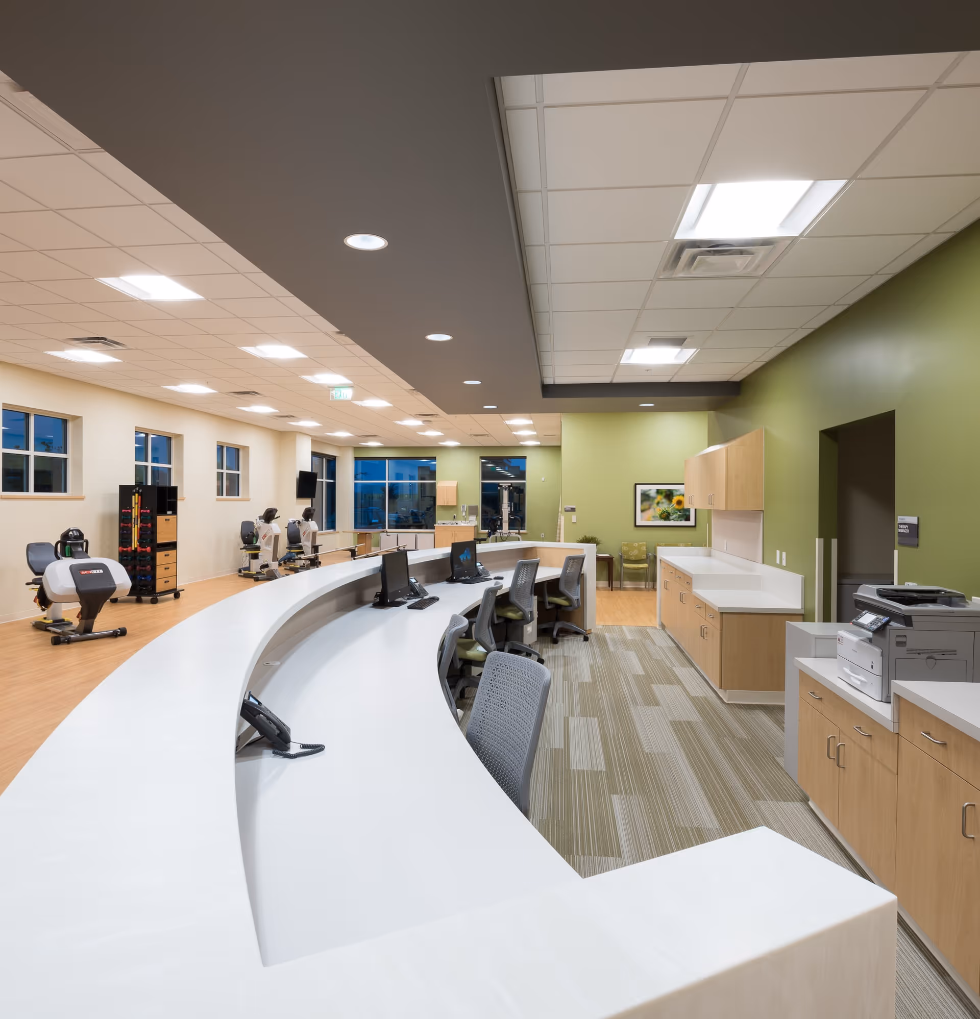 Interior view of a modern reception or nurse station area in a senior living facility with a curved white desk, office chairs, computer monitors, telephones, and cabinetry. In the background, there is exercise equipment and large windows letting in natural light. The walls are painted green and beige, and the ceiling has recessed lighting.