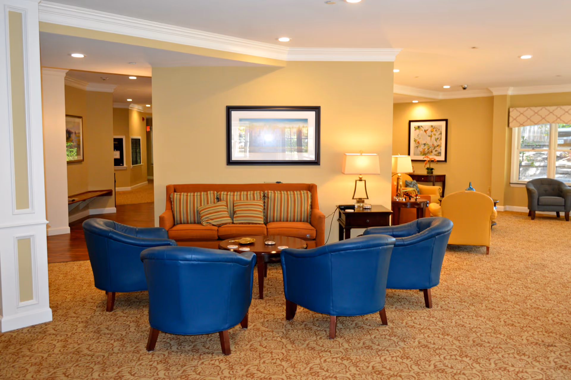 A cozy seating area in a senior living facility with a brown sofa adorned with striped cushions, surrounded by four blue armchairs arranged around a wooden coffee table. The room has beige walls, carpeted floors, framed artwork, and table lamps providing warm lighting. Additional seating and windows are visible in the background.