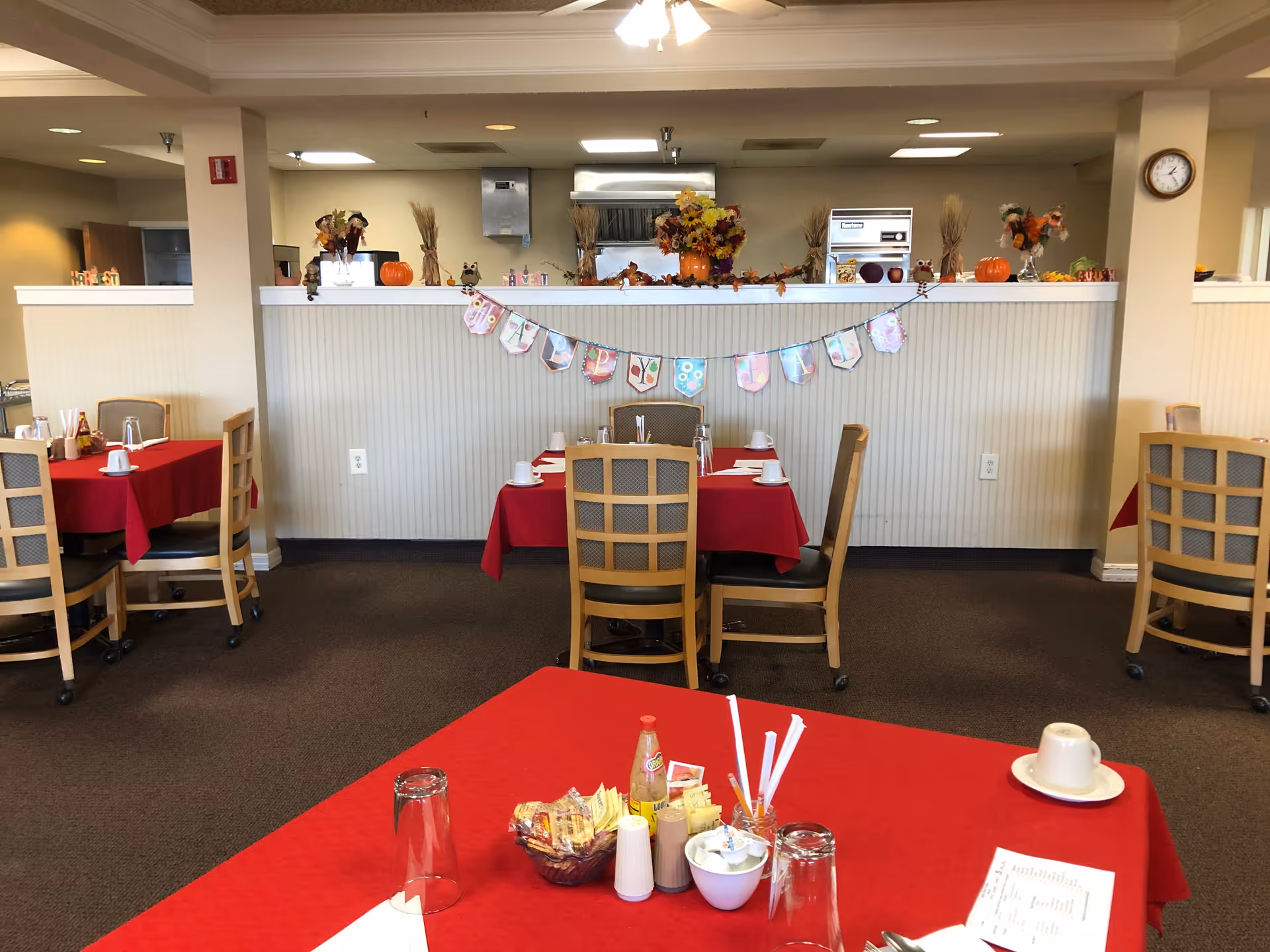 Dining room with several tables covered in red tablecloths, each set with cups, glasses, and utensils. The room has beige walls and a partial divider decorated with fall-themed items including pumpkins, flowers, and a banner that reads 'Happy Fall'. Chairs with wooden frames and cushioned seats surround the tables. A clock is visible on the wall to the right.