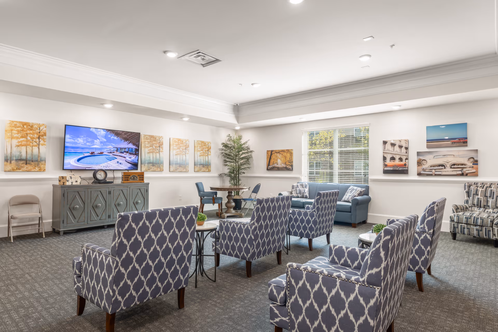 Bright communal lounge with patterned armchairs and sofas arranged around side tables, a TV on a console, and wall artwork.
