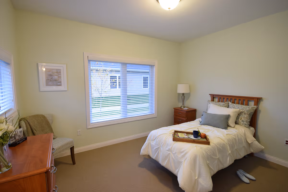 A cozy bedroom with a single wooden bed dressed in white bedding and multiple pillows. A wooden nightstand with a lamp is beside the bed. A wooden dresser with a vase of flowers and a chair with a green throw blanket are against the opposite wall. A large window with blinds lets in natural light, showing a view of the outside. A pair of slippers is placed on the floor near the bed.