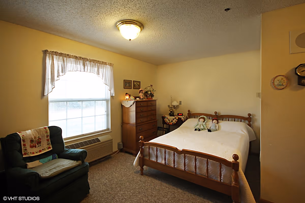 A modest bedroom with a wooden bed topped by two dolls, a dresser, and an upholstered armchair by a window.