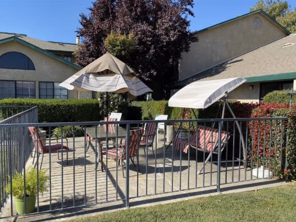 Outdoor patio area at Sutter Estates Retirement Community with a metal railing surrounding a concrete floor. The patio has a table with several chairs featuring red and white striped cushions, two large umbrellas providing shade, and a cushioned swing seat with a white canopy. The background shows a hedge, trees, and parts of the building with beige walls and green trim under a clear blue sky.