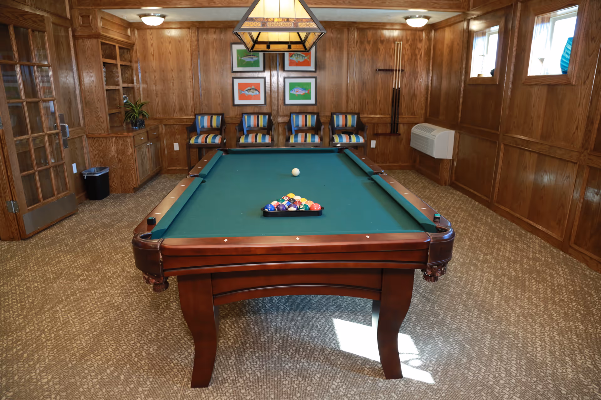 A room with wood-paneled walls and carpeted floor featuring a billiards table with a full rack of pool balls and a cue ball. Four striped cushioned chairs are lined up against the far wall beneath four colorful fish paintings. A hanging light fixture is positioned above the pool table. There is a wooden door with glass panes on the left and a wall-mounted air conditioning unit on the right.