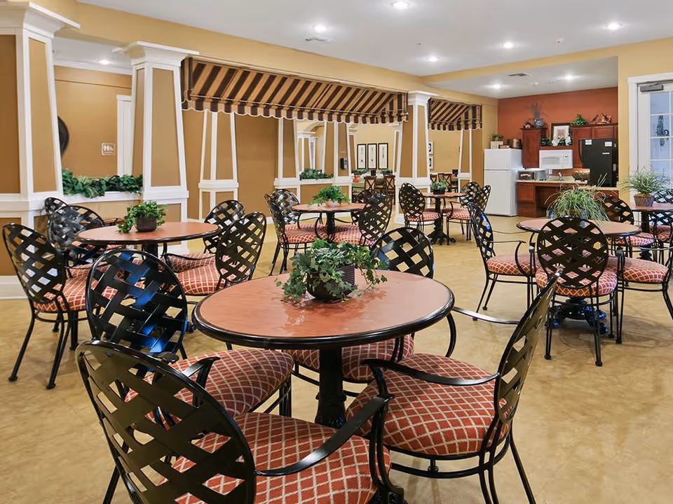 A dining area with multiple round tables and black metal chairs with red patterned cushions. Each table has a small green plant centerpiece. The room has beige walls with white trim, a striped brown and beige awning over a serving counter, and a kitchen area with a refrigerator, microwave, and cabinets in the background.